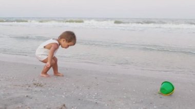 Çocuk yaz güneşinin altında kumlu bir zeminde deniz kabukları ve çakıl taşları topluyor.