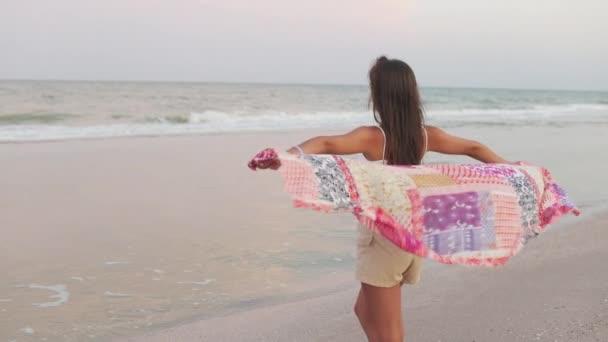 Dziewczyna w letniej koszulce i spodenkach spaceruje po plaży patrząc na horyzont i trzymając szal w rękach. — Wideo stockowe