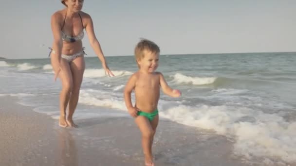 Девушка в купальнике бегает с малышом на пляже с раковинами на фоне моря с волнами — стоковое видео