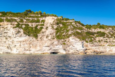 Light-colored layered rock creating cave on beach of Corfu island in Greece with clear turquoise water under blue cloudless sky