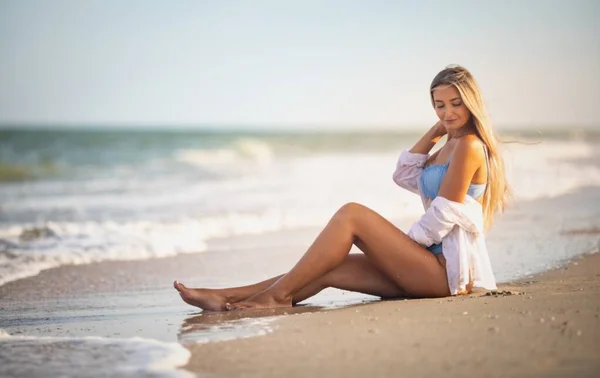 Egy lány kékes fürdőruhában és ingben ül a vízparton, és belenéz a horizontba. — Stock Fotó