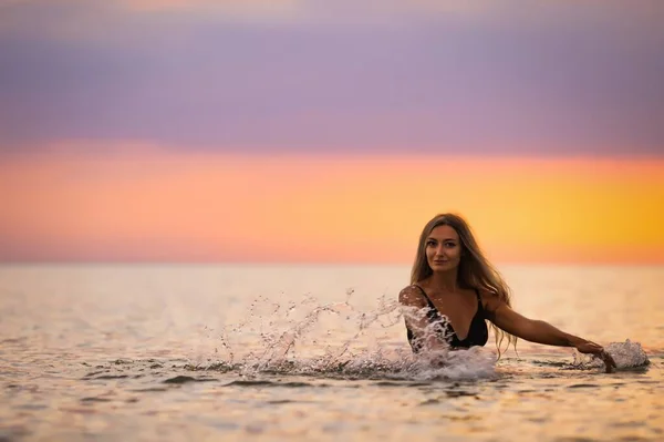 A girl with blond hair in a black swimsuit splashes to the sides while sitting in an estuary on a sunset background — Stock Photo, Image