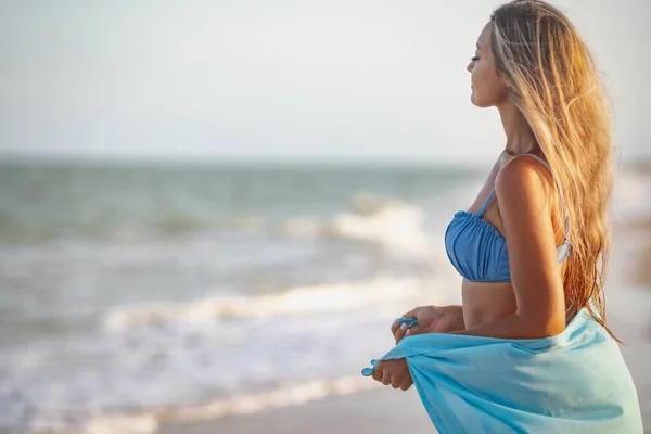 A girl with blond hair in a bluish swimsuit and a bright shawl walks along the beach — Stock Photo, Image