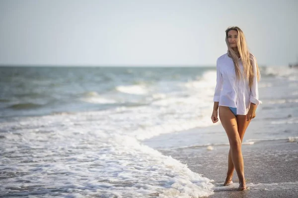 Uma menina delgada em um terno de banho azul suave e camisa caminha ao longo da praia de areia perto do mar azul — Fotografia de Stock