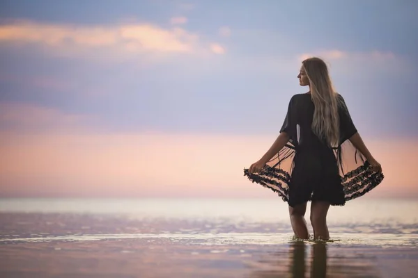 Uma menina com cabelo loiro chique em um biquíni preto e um xale de renda caminha ao longo do estuário — Fotografia de Stock