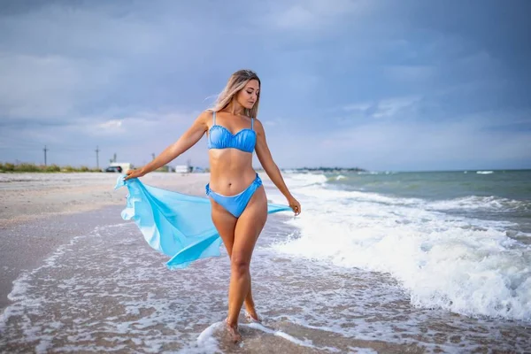 Uma menina com cabelo loiro em um maiô azulado e um xale brilhante caminha ao longo da praia — Fotografia de Stock