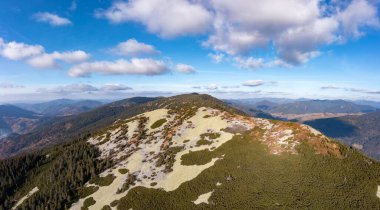 Güneşli bir günde mavi gökyüzünde süzülen beyaz kabarık bulutların altında, yamaçlardaki kozalaklı ormanlarla ve ormansız alanlarla yüksek dağ zirvesi