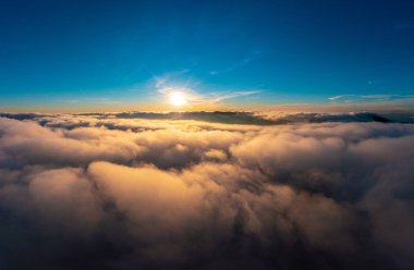 Sun light shining brightly on blue sky above thick layer of white fluffy clouds at sunrise with mountains on background panorama view