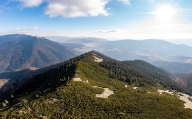 Güneşli bir günde mavi gökyüzünde süzülen beyaz kabarık bulutların altında, yamaçlardaki kozalaklı ormanlarla ve ormansız alanlarla yüksek dağ zirvesi