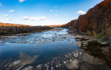 Kayalık dibi olan sığ dağ nehri ve sonbahar günü mavi bulutsuz gökyüzünün altında tepedeki kozalaklı terasotta ormanları.