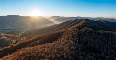 Sonbahar terakotta ormanı ve mavi bulutsuz gökyüzü manzarası altında küçük evler ile yüksek dağ zirvelerinde parlayan gündoğumu