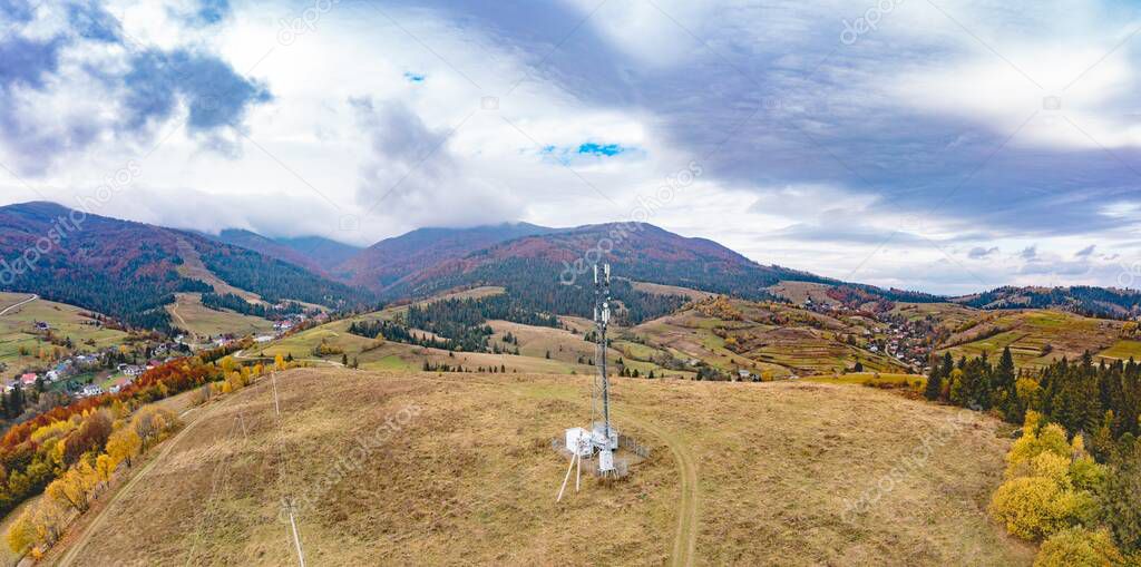 High steel cell tower near settlement on mountain top covered with autumn forest under gloomy sky with thick grey clouds panorama view