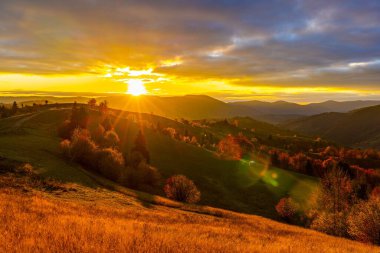 Yükseklerde yemyeşil ağaçlarla dolu çimenli yamaçlar. Sonbaharda bulutlu gökyüzünün altında günbatımında uzak dev orman dağlarına karşı.
