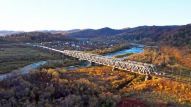 Dağlık arazide renkli ağaçlarla dolu bir nehir üzerindeki demiryolu köprüsü.