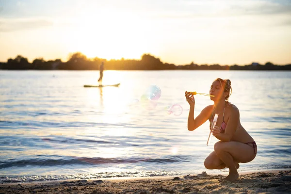 Mulher em um maiô soprando bolhas de sabão andando ao longo da praia perto do lago em férias de verão — Fotografia de Stock