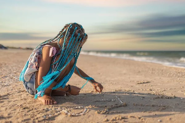 Egy afrikai hajú lány nyári jelmezben játszik a tengerparton kagylókkal a tenger közelében, napnyugta alatti hullámokkal. — Stock Fotó