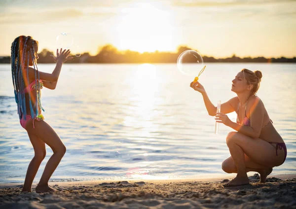 Mãe brinca com sua filha soprando bolhas na costa perto do lago — Fotografia de Stock