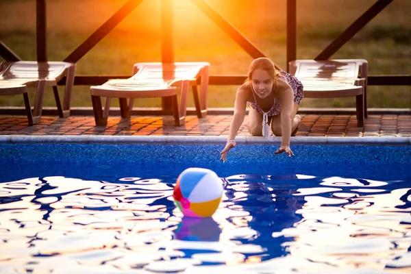 Una chica con el pelo trenzado en un moño en traje brillante juega junto a la piscina con una pelota contra el fondo del sol de verano — Foto de Stock