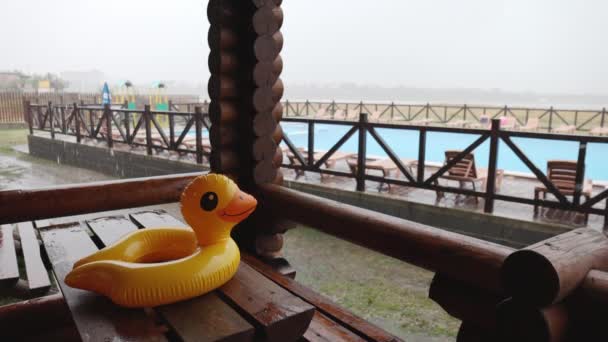 An inflatable duck lies on wooden table in gazebo at recreation center against the backdrop of pool covered with rain — Stock Video