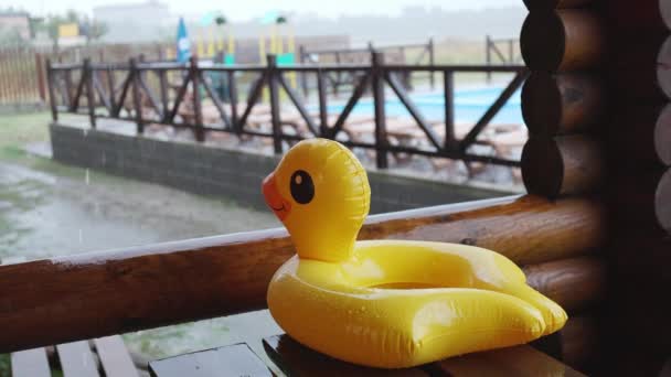 Un canard gonflable se trouve sur une table en bois dans un kiosque au centre de loisirs, sur fond de piscine couverte de pluie — Video