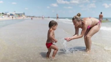 Anne, yaz güneşinin altında tatil yaparken denizde durup gülen bebeğinin üzerine su sıçratır.
