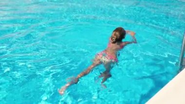 Mor gözlüklü pembe mayo giyen bir kız yaz günü mavi suyla dolu bir havuzda yüzer.