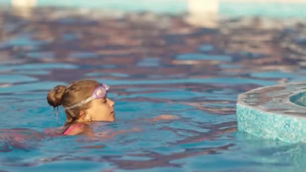 En flicka i en ljus baddräkt med simglasögon dyker ner i en pool med klart genomskinligt vatten — Stockvideo