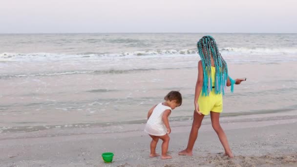 Мальчик в полосатой футболке танцует под музыку со своей сестрой африканскими косичками на пляже у моря — стоковое видео