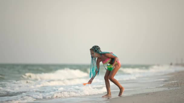 Egy afrikai hajú lány nyári jelmezben játszik a tengerparton kagylókkal a tenger közelében, napnyugta alatti hullámokkal. — Stock videók