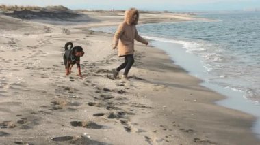 Ceketli bir kız sahilde Rottweiler köpeğiyle koşuşturuyor.