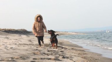 Ceketli bir kız sahilde Rottweiler köpeğiyle koşuşturuyor.