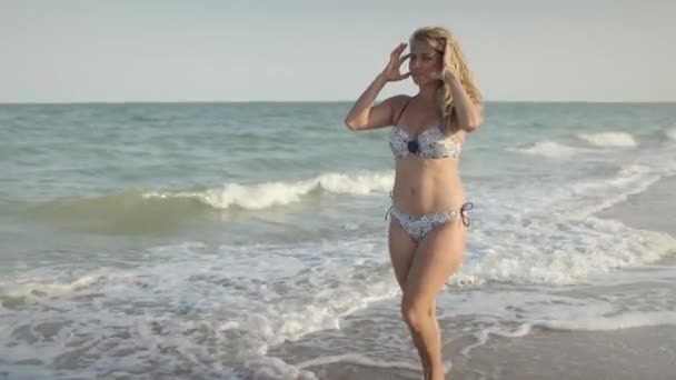 En flicka i en ljus baddräkt går längs stranden med snäckor nära havet med vågor — Stockvideo