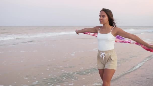 Девушка в летней футболке и шортах идет по пляжу, глядя на горизонт и держа в руках шаль — стоковое видео