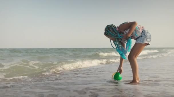 En flicka med afrikanska flätor i en sommarkostym leker på stranden med skal nära havet med vågor under solig solnedgång — Stockvideo