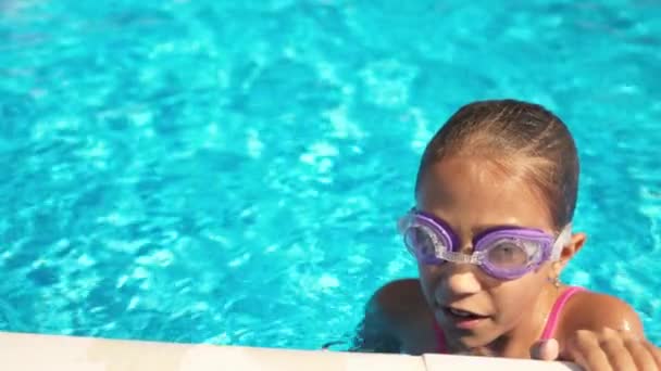 Egy rózsaszín fürdőruhás lány lila szemüvegben lebeg a medencében kék vízzel egy nyári napon. — Stock videók