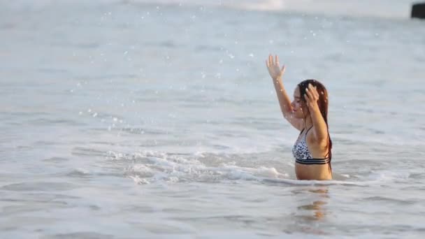 Nedves hajú lány leopárdos fürdőruhában vizet fröcsköl, miközben a tengerben ül. — Stock videók