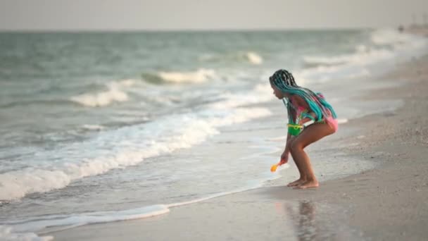 Ein Mädchen mit afrikanischen Zöpfen im Sommerkostüm spielt am Strand mit Muscheln am Meer mit Wellen unter sonnigem Sonnenuntergang — Stockvideo