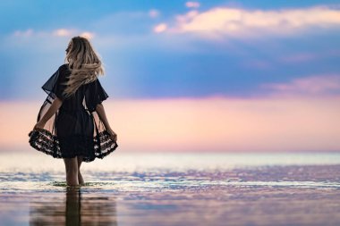 Siyah bikini mayo ve siyah dantelli şalla sarışın, ince, güzel bir kız sığ bir nehir kenarında, güneşli bir yaz günbatımının arka planında yürüyor.