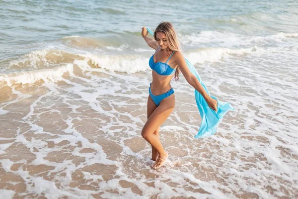優しいブルーの水着と明るいライトブルーのショールでブロンドの髪の若い細い日焼けした女の子は 暖かい夏を楽しんで海の水の端近くのビーチに沿って歩きます — ストック写真
