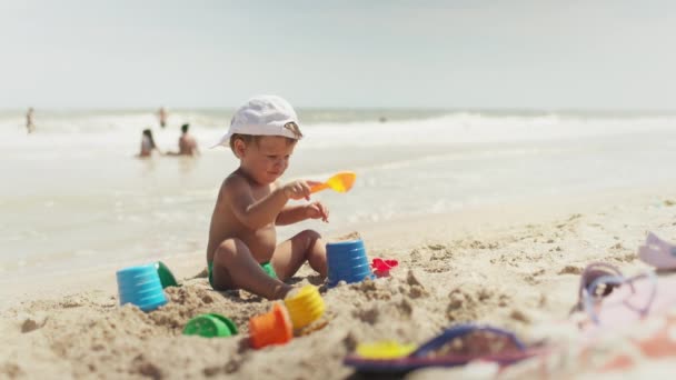 Chico jugando con juguetes en la playa construyendo perlas y torretas sonriendo a alguien detrás de las escenas en las vacaciones de verano — Vídeos de Stock