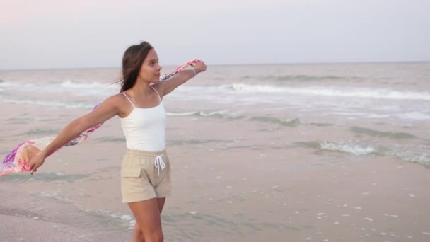 Дівчина в літній футболці і шортах ходить уздовж пляжу, дивлячись на горизонт і тримаючи в руках хустку — стокове відео