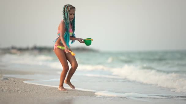 En flicka med afrikanska flätor i en sommarkostym leker på stranden med skal nära havet med vågor under solig solnedgång — Stockvideo