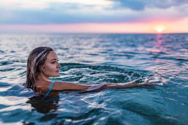 Bronz tenli bir kız ıslak sarı saçlı, gün batımının arka planında şeffaf suyla temiz bir nehir kenarında otururken bilinmeyen bir mesafeye bakıyor.