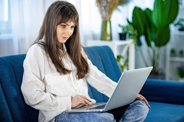 La chica está sentada en el sofá y trabajando en un ordenador portátil en el apartamento — Foto de Stock