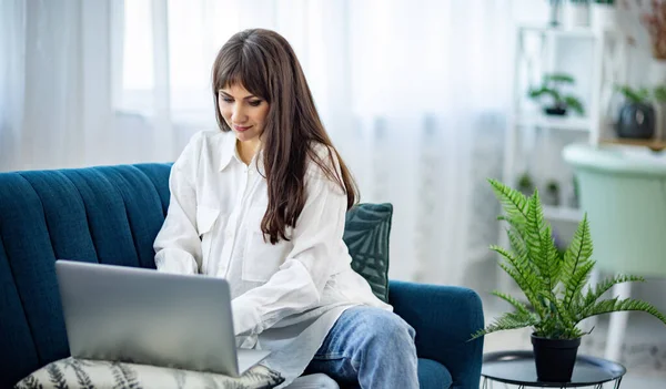 La chica está sentada en el sofá y trabajando en un ordenador portátil en el apartamento — Foto de Stock