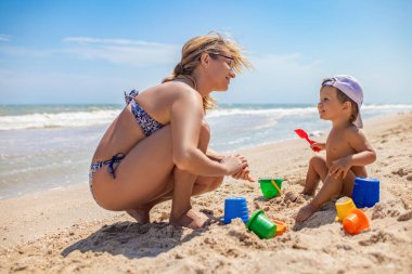 Gözlüklü anne, deniz kenarında oturan küçük bir bebekle oynuyor.