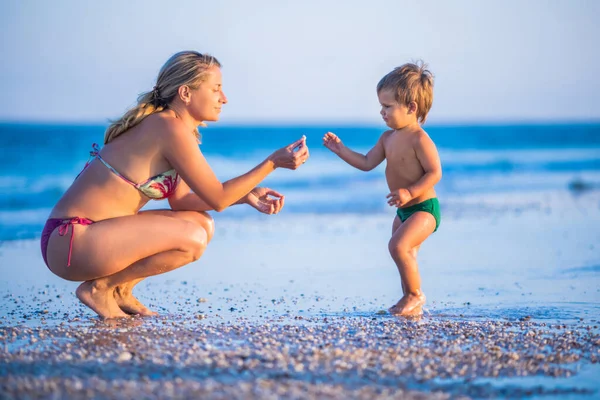 Mamma con gli occhiali gioca con un piccolo bambino seduto sulla spiaggia vicino al mare — Foto Stock