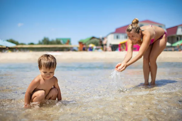 Mamá juega y salpica en su bebé que se ríe mientras está de pie en el mar bajo el sol de verano de vacaciones — Foto de Stock