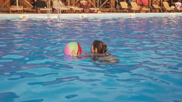 Una chica en un traje de baño brillante nada con una pelota inflable en una piscina con agua clara en una noche de verano — Vídeos de Stock