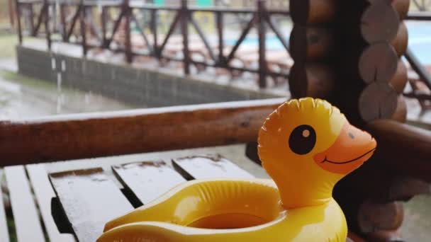 Um pato inflável encontra-se na mesa de madeira no gazebo no centro de recreação contra o pano de fundo da piscina coberta com chuva — Vídeo de Stock